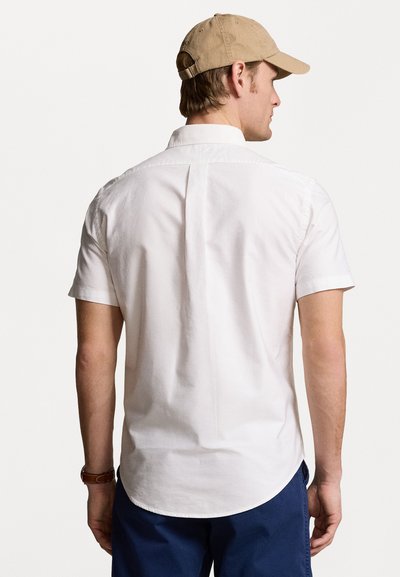 White short-sleeve shirt with a straight hem and back pleat, paired with dark blue pants and a beige baseball cap.