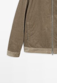 Giacca in corduroy marrone con rigatura verticale, chiusura con zip, tasche laterali e un tessuto più chiaro a contrasto all'orlo e ai polsini.