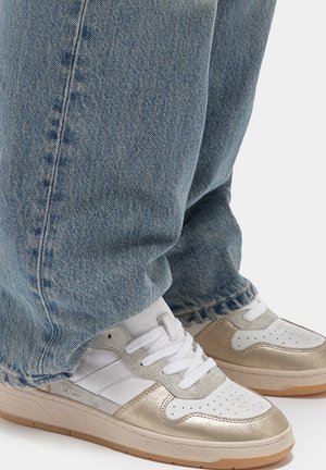 Sneaker bianchi e dorate con punta perforata, lacci sul davanti e suola in gomma marrone chiaro; abbinate a jeans di denim azzurro.