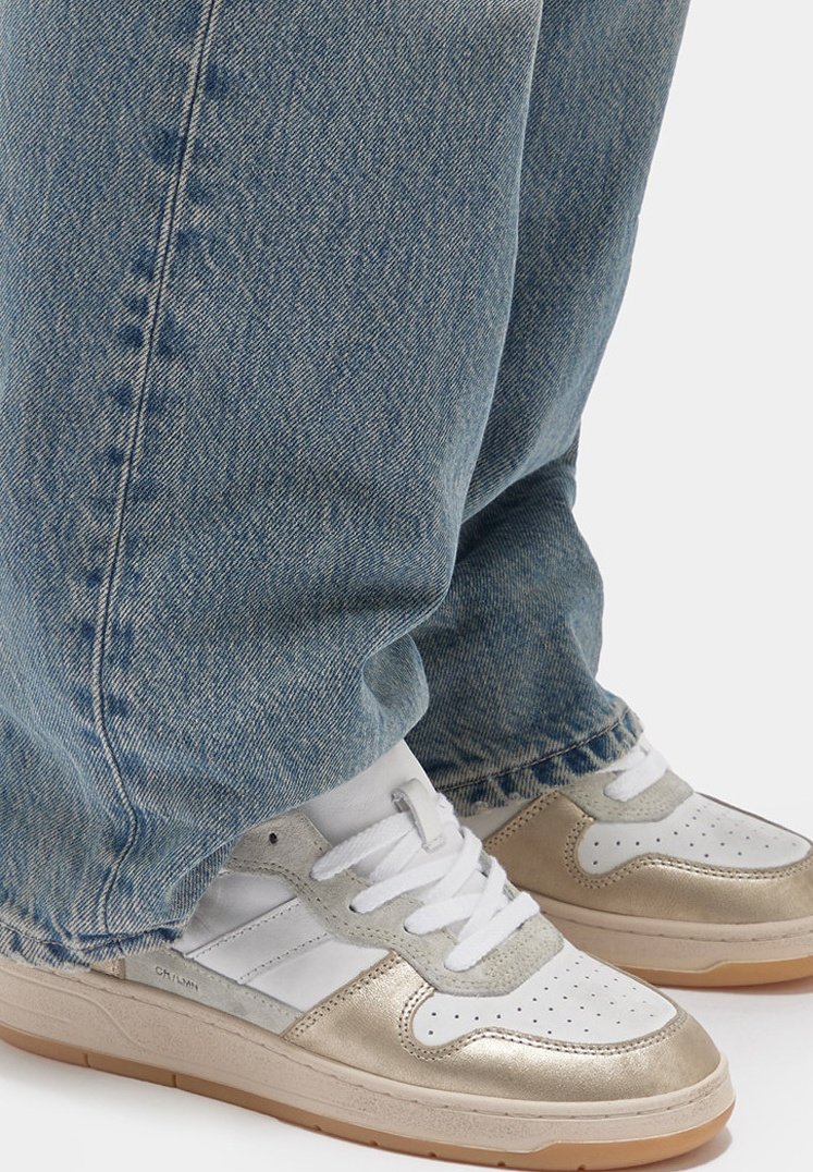 Sneaker bianchi e dorate con punta perforata, lacci sul davanti e suola in gomma marrone chiaro; abbinate a jeans di denim azzurro.