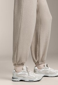 Világos bézs pamut melegítőnadrág elasztikus mandzsettákkal, fehér sportcipőkkel párosítva, amelyek szürke kiemelésekkel és texturált rétegekkel rendelkeznek. Összetételük alkalmassá teszi őket hétköznapi viseletre.