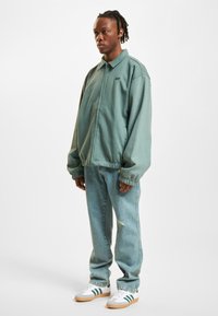Oversized, soft green jacket with a zip front, ribbed cuffs, and collar. Paired with light blue jeans and gray sneakers with green stripes.