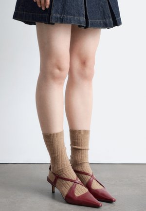 Jambes portant une jupe en jean foncé, des chaussettes côtelées marron et des chaussures bordeaux à talon bas avec des lanières croisées, debout sur un sol gris.