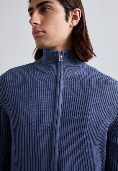 Ναυτικό πλεκτό πουλόβερ με ψηλό γιακά και κλειστό φερμουάρ κατά το 1/4. Ο δομημένος ύφασμα δημιουργεί κατακόρυφες σχέσεις στην επιφάνεια του ρούχου.