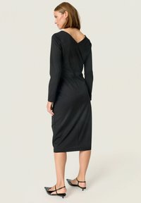 Vestido negro ajustado con mangas largas, escote en la espalda y corte a media longitud. Textura suave y silueta simple, combinado con tacones negros de tiras.