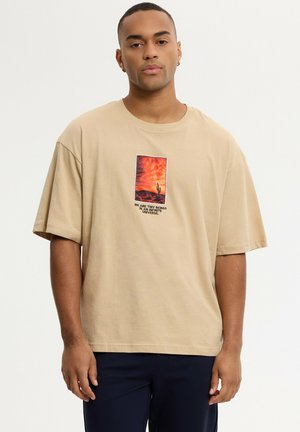 Joven con una camiseta beige con estampado de un desierto al atardecer y el texto "We are tiny beings in an infinite universe."