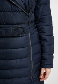 Cabană puffer navy cu textură matlasată, având un fermoar frontal, o curea ajustabilă cu o cataramă circulară și buzunare laterale.