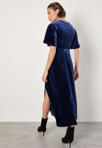 Robe maxi en velours bleu marine avec des manches courtes et larges et des fentes sur les côtés. Portée avec des bottines noires, offrant une texture lisse et une silhouette élégante.