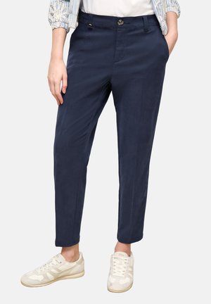 Person trägt marineblaue, schmal zulaufende Hose mit vorderer Knopfleiste und weißen Schnürsneakern, Hände in den Taschen, vor einfarbigem Hintergrund.