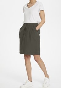 Jupe en coton vert avec une taille élastique, deux poches latérales, design longueur genou et cordon de serrage ; associée à un t-shirt blanc et des baskets blanches.