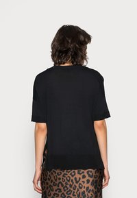 Kobieta z krótkimi kręconymi włosami, ubrana w luźną czarną bluzkę z krótkim rękawem i brązowo-czarną spódnicę w panterkę, zwrócona tyłem.