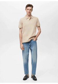 Beige polo-shirt, lige ben denim jeans i lys blå og sorte slip-on sko. Modellen står med hænderne i lommerne foran en ensfarvet baggrund.