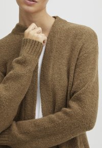 Bruine gebreide cardigan met een ontspannen pasvorm, voorzien van geribde manchetten en een textuur die zacht en warm lijkt. Draagt over een witte top.