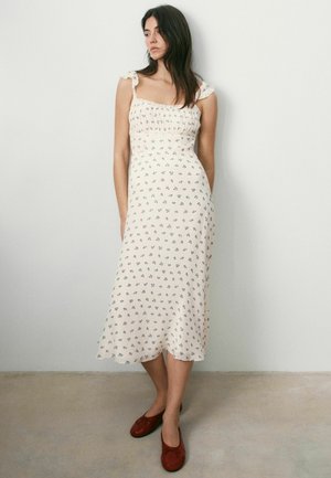 Woman in a white midi dress with small floral print and tied shoulder straps, wearing red flat shoes, standing against a plain white wall.