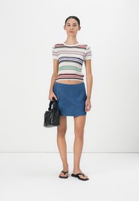 Gestreiftes Strickoberteil in verschiedenen Farben, Denim-Minirock, schwarze strukturierte Handtasche und schwarze Flip-Flops; Modell steht vor einem weißen Hintergrund.