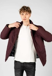 Giacca isolante bordeaux con chiusura a zip e bottoni a pressione, con fodera interna grigia. Il modello indossa una t-shirt bianca e jeans scuri.