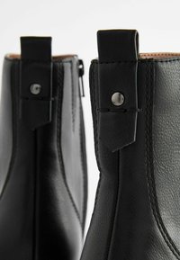Stivaletti alla caviglia in pelle nera con texture liscia, dettagli cuciti e accenti metallici sui talloni. Chiusura con zip laterale visibile.