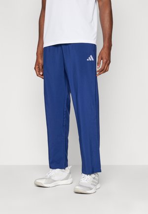 Mężczyzna ubrany w niebieskie sportowe spodnie Adidas, białe trampki i białą koszulę, stoi na tle jasnego, jednolitego tła.