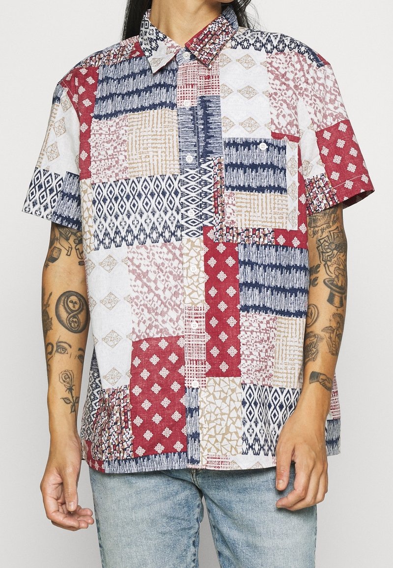 Chemise patchwork à manches courtes présentant des motifs variés en rouge, blanc, bleu et beige, avec un col à revers et une poche avant.