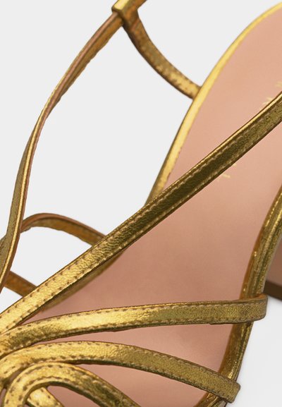 Κοντινή λήψη χρυσού μεταλλικού πέδιλου με λουράκια, υφή στο φινίρισμα και απαλό ροζ πάτο, πάνω σε ανοιχτό φόντο.