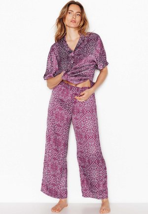 Vrouw draagt een paars met patroon kortarmig shirt en bijpassende wijde broek, staat blootsvoets tegen een witte achtergrond.