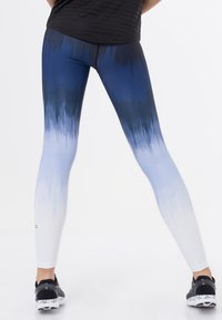 Osoba oblečená v černých teniskách a sportovních legínách s modro-bílým plynulým přechodem, stojící s roztaženýma nohama na jednobarevném bílém pozadí.