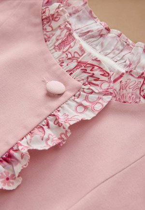 Prim-plan al unui articol vestimentar roz, cu o nasture acoperit cu țesătură și un volan cu imprimeu floral alb și roz în jurul gâtului.
