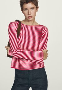Vrouw met kort haar, gekleed in een rode en witte gestreepte lange mouwtop en donkere broek, met een bruine schoudertas tegen een eenvoudige achtergrond.