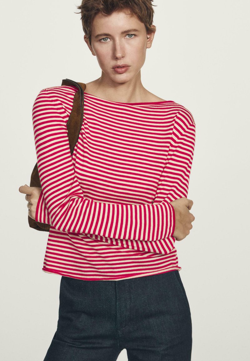 Vrouw met kort haar, gekleed in een rode en witte gestreepte lange mouwtop en donkere broek, met een bruine schoudertas tegen een eenvoudige achtergrond.