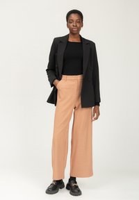 Schwarzer, doppelt geknöpfter Blazer über einem schwarzen gerippten Oberteil, kombiniert mit helloranfarbenen, weiten Hosen und schwarzen, klobigen Loafers mit goldenen Akzenten.