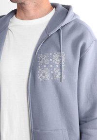 Hoodie zippé bleu clair avec une poche frontale, présentant un design floral blanc et du texte sur la partie supérieure gauche de la poitrine, fabriqué en tissu doux.