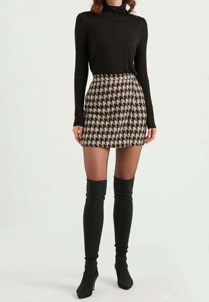 Mujer con un suéter negro de manga larga y cuello alto, falda mini de pata de gallo en beige y negro, medias transparentes y botas altas de tacón negras, de pie.