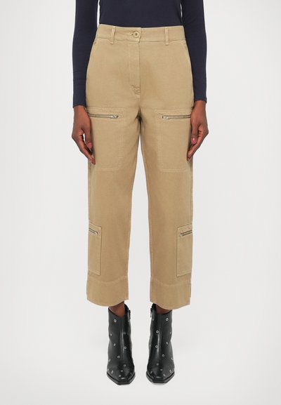 Pantaloni cargo beige con due tasche anteriori con zip, toppe laterali e lunghezza cropped. Indossati con stivali neri con tacco alla caviglia caratterizzati da accessori metallici.