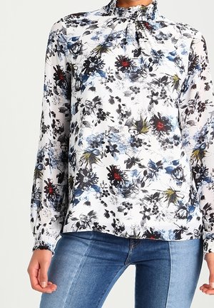 Vrouw draagt een witte blouse met lange mouwen met zwarte, blauwe, rode en gele bloemmotieven en een blauwe spijkerbroek.