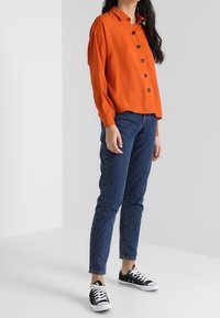 Orange button-up skjorte med lange ærmer, parret med mørkeblå denim jeans og sorte og hvide sneakers. Enkel design og klassisk pasform.