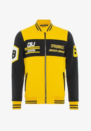 Geel en zwarte zip-up jack met gestreepte accenten, logo patches en twee zijzakken. Draagt het met blauwe jeans en zwarte sportschoenen.