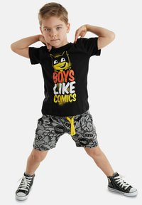 Sort t-shirt med grafisk comicfigur og teksten "DRENGE KAN LIDE COMICS". Grå shorts med dristige comic-strip mønstre og gul snor. Sorte sneakers.