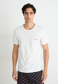 Weisses Baumwoll-T-Shirt mit kurzen Ärmeln, das auf der linken Brust ein kleines rotes "HUGO"-Logo zeigt. Einfacher runder Halsausschnitt.