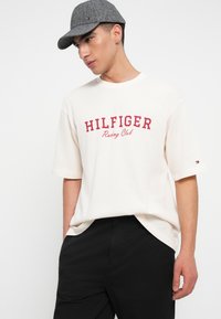 Νέος άνδρας φορώντας ένα γκρι καπέλο, κρεμ κοντομάνικο πουκάμισο με κόκκινο κείμενο "Hilfiger Racing Club" και μαύρο παντελόνι, ποζάροντας με τα χέρια στα τσέπες.