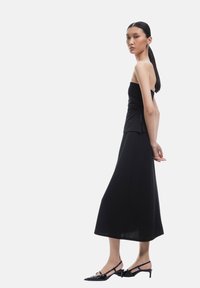 Haut sans bretelles noir associé à une longue jupe noire, fabriquée dans un tissu lisse. Présente une fente sur le côté et des talons aiguilles avec des nœuds.