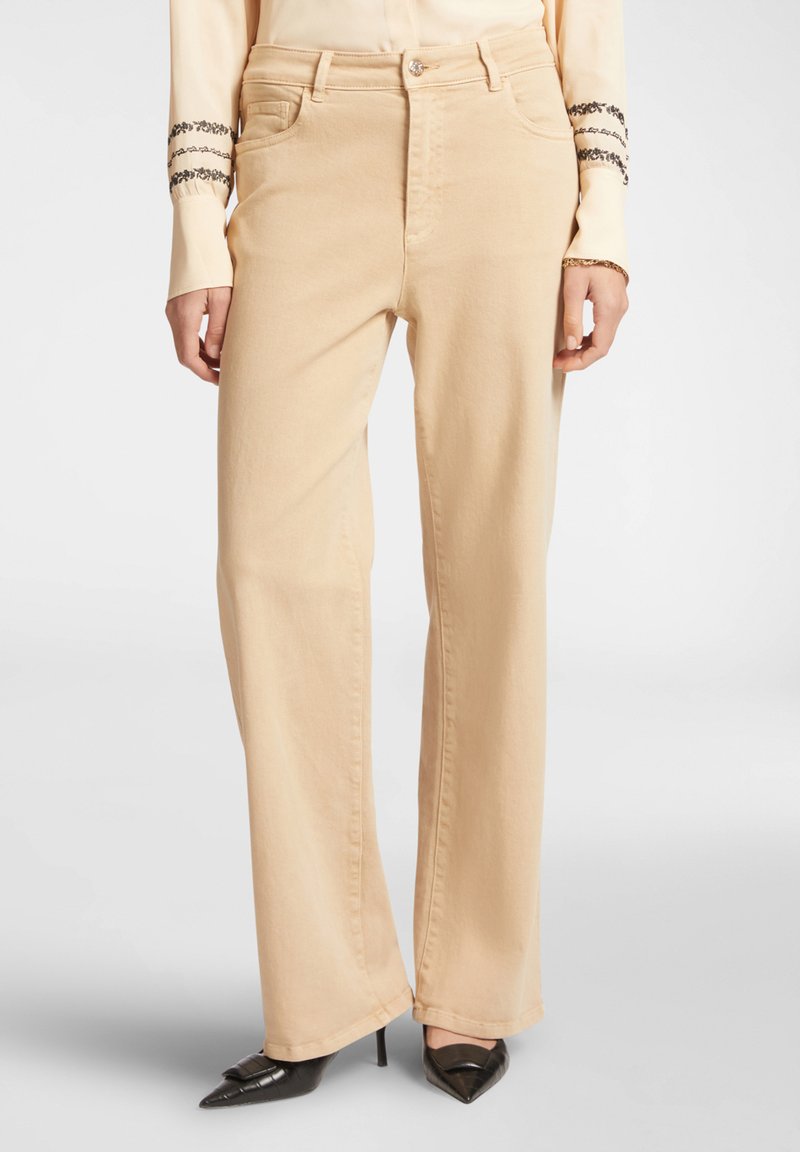 Pantaloni beige a gamba larga con una texture liscia, dotati di passanti per cintura, tasche frontali e abbinati a décolleté nere a punta.