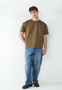 T-shirt en coton marron à manches courtes, associé à un jean en denim bleu clair ample et des chaussures noires. Le mannequin se tient devant un fond blanc.