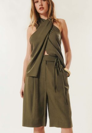 Femme portant un haut cache-cœur sans manches vert olive et un short taille haute assorti, debout avec une main dans la poche sur un fond neutre.