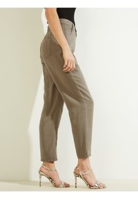 Pantalones caquis ajustados con bolsillos laterales y un bolsillo trasero que presenta un pequeño logo, combinados con sandalias de tacón alto metálicas con tiras.