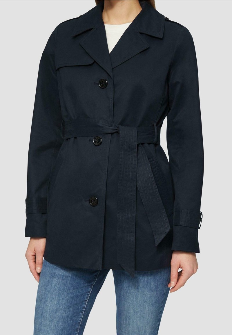 Femme portant un trench-coat bleu marine foncé ceinturé avec des boutons, associé à un jean bleu et un haut blanc en dessous.