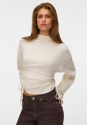Femme aux longs cheveux portant un haut blanc froncé à manches longues et un jean taille haute foncé sur un fond blanc uni.