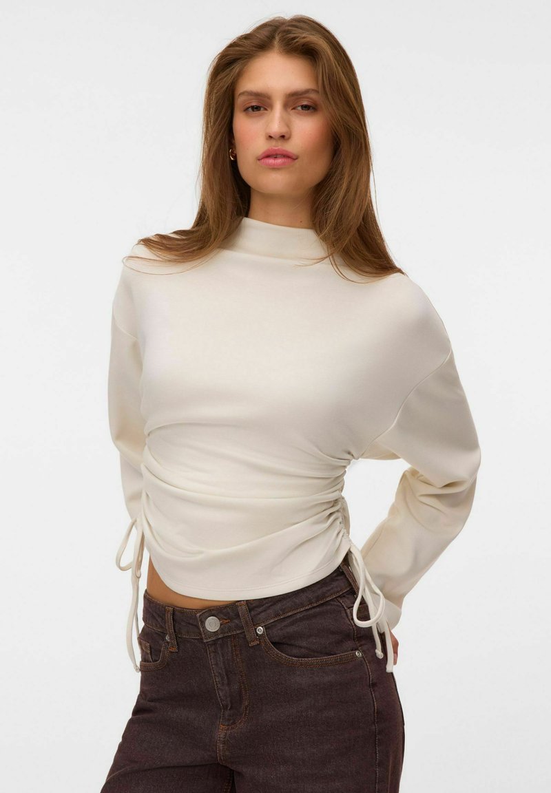Femme aux longs cheveux portant un haut blanc froncé à manches longues et un jean taille haute foncé sur un fond blanc uni.