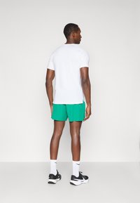 Weißes kurzärmeliges Hemd, kombiniert mit grünen Shorts, die ein figurbetontes Design und seitliche Schlitze aufweisen. Getragen mit schwarzen Turnschuhen und weißen Socken.