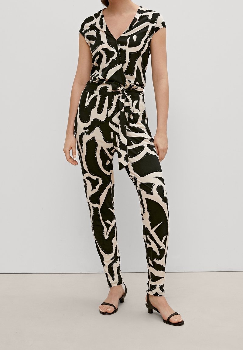 Femme portant un haut cache-cœur à motif abstrait noir et beige et un pantalon assorti noué à la taille avec des talons ouverts noirs, debout contre un mur uni.