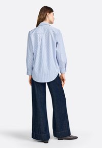 Camicia a righe blu chiaro e bianco con bottoni, maniche lunghe, yoke posteriore e orlo curvo, abbinata a pantaloni in denim scuri a gamba larga.